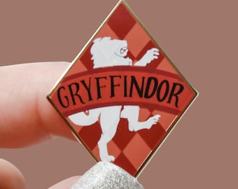 Könnte beinhalten: Ein rautenförmiger Ansteckpin mit dem Wappen des Hauses Gryffindor aus der Harry-Potter-Reihe. Der Pin hat ein rot-goldenes Farbschema mit einem weißen Löwenemblem und dem Wort "GRYFFINDOR" in einem geschwungenen Banner.
