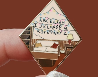 Könnte beinhalten: Ein rautenförmiger Emaille-Pin mit goldenem Rand, der eine Wohnzimmerszene mit Sofa, Lampe und Buchstabenlichtern darstellt. Der cremefarbene Hintergrund des Pins zeigt das Alphabet von A bis Z.