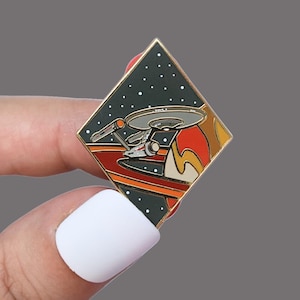 Pode incluir: Um alfinete de esmalte em forma de losango com a nave espacial Star Trek Enterprise em um fundo de um planeta e céu estrelado. O alfinete tem uma borda dourada e é segurado por uma pessoa.