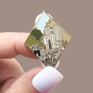 May include: A diamond-shaped enamel pin featuring a detailed landscape with a white castle, green hills, and a light blue sky. The pin has a gold-colored border and is held by a person.