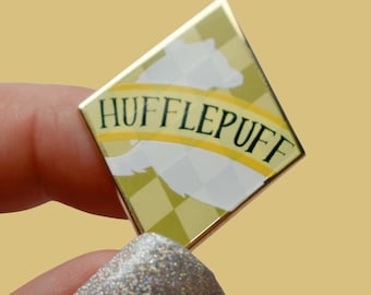 Könnte beinhalten: Eine goldfarbene, rautenförmige Anstecknadel mit dem Text "HUFFLEPUFF" in schwarzer Schrift. Die Anstecknadel zeigt eine weiße Dachs-Silhouette auf einem gelb-weiß karierten Hintergrund. Die Anstecknadel wird von einer Person gehalten.