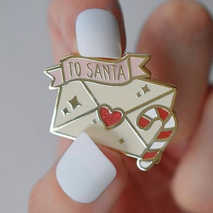 Pode incluir: Um pin de esmalte dourado em forma de carta para o Papai Noel. O pin apresenta um envelope creme com um coração vermelho brilhante, uma bengala doce e uma faixa rosa que diz "TO SANTA".