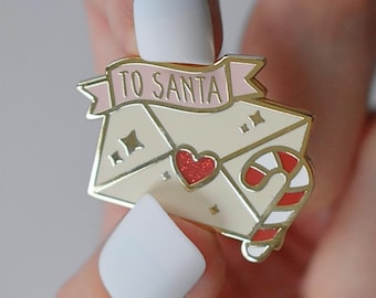 May include: A gold-toned enamel pin shaped like a letter to Santa. The pin shows a cream-coloured envelope with a red glitter heart, a candy cane, and a pink banner that reads "TO SANTA".