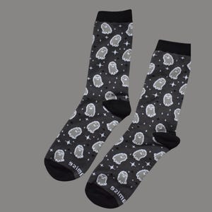 Puede incluir: Un par de calcetines grises oscuros con un patrón repetido de fantasmas y estrellas blancas. Los calcetines tienen talones y dedos negros, y el texto "92ime" está impreso en la parte inferior de cada calcetín.