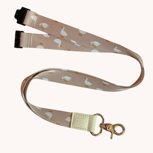 May include: A dusty pink lanyard featuring a repeating pattern of small white geese with orange beaks and feet. The fabric strap includes two black plastic connectors at the top and a gold-toned metal lobster clasp attached to a cream-coloured leather tab.