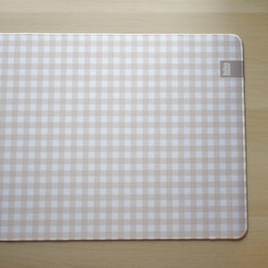 May include: A rectangular beige and white gingham pattern placemat with rounded corners. The placemat has a small rectangular tag in the upper right corner. The placemat is on a light brown wooden surface.