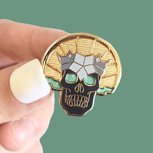 May include: A black and gold enamel pin featuring a skull wearing a crown with a geometric design. The skull has green eyes and a gold outline. The crown is silver with a white geometric design.