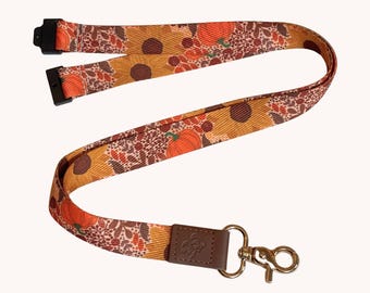 Könnte beinhalten: Ein Schlüsselband aus Stoff mit einem herbstlichen Muster aus orangefarbenen Kürbissen, gelben Sonnenblumen und braunem Laub. Das Band verfügt über schwarze Kunststoffverschlüsse oben und eine braune Kunstlederlasche an einem silberfarbenen Karabinerhaken.