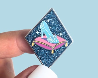 Könnte beinhalten: Eine rautenförmige Emaille-Anstecknadel mit glitzerndem dunkelblauem Hintergrund und weißen Sternakzenten. In der Mitte befindet sich ein hellblauer Stöckelschuh auf einem rosa Kissen mit gelben Quasten an jeder Ecke.