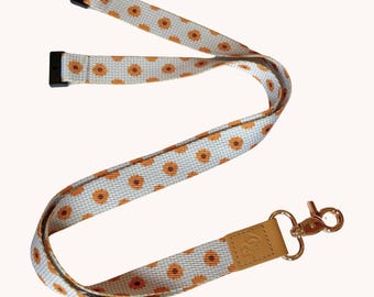 Könnte beinhalten: Ein Schlüsselband aus Stoff mit einem sich wiederholenden Muster aus orangefarbenen und braunen Sonnenblumen auf hellblauem Grund. Das Band hat zwei schwarze Kunststoff-Sicherheitsverschlüsse oben und eine beigefarbene Lasche mit einem goldenen Karabinerhaken.