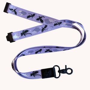 May include: A light purple fabric lanyard featuring a repeating pattern of black dragon silhouettes, white crescent moons, and small white stars. The lanyard includes black plastic hardware, a black leather-like connector, and a black metal lobster clasp.