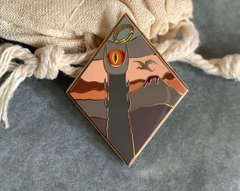 May include: A diamond-shaped enamel pin featuring a tower with an orange eye, a ring, and a bird silhouette against a sunset backdrop. The pin has a gold border and is set against a neutral fabric.