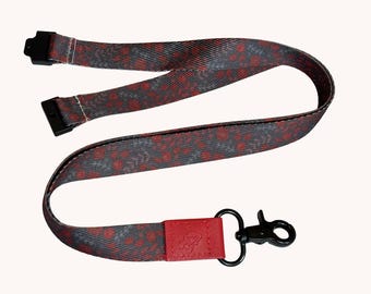 Könnte beinhalten: Ein Schlüsselband aus Stoff mit einem dunkelgrauen und roten Blumenmuster. Das Band verfügt über schwarze Kunststoffanschlüsse an den Enden und eine rote lederartige Lasche, die an einem schwarzen Metallkarabiner zur Befestigung befestigt ist.