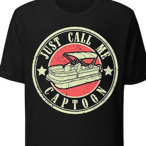 May include: Black t-shirt featuring a circular graphic with a pontoon boat illustration. The graphic includes the text "JUST CALL ME CAPTOON" in a distressed, vintage style. The design is in shades of red, beige, and green.
