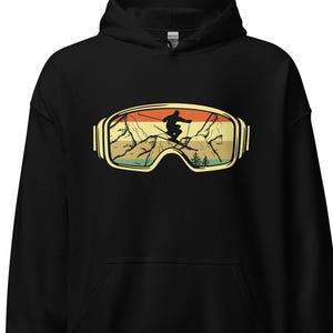 May include: Black hoodie with a graphic of ski goggles, a skier silhouette, and a mountain scene. The goggles are gold-coloured, and the design uses a retro colour scheme of orange, green, and yellow.