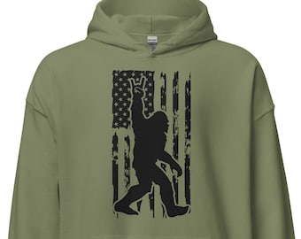 Peut inclure: Un sweat-shirt à capuche vert avec une silhouette noire de Bigfoot faisant un geste de rock and roll devant un drapeau américain.