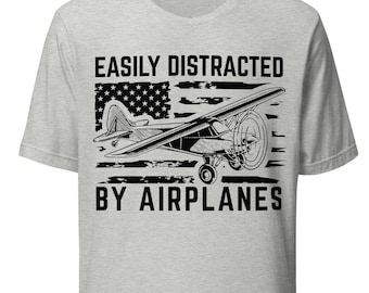 May include: Light gray t-shirt with a black and white graphic of an airplane and the American flag. The text reads "EASILY DISTRACTED BY AIRPLANES". The shirt is made of a soft material.