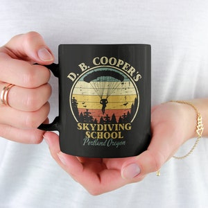 May include: Black ceramic mug with a vintage-style design featuring a skydiver and the text "D.B. Cooper's Skydiving School Portland Oregon".
