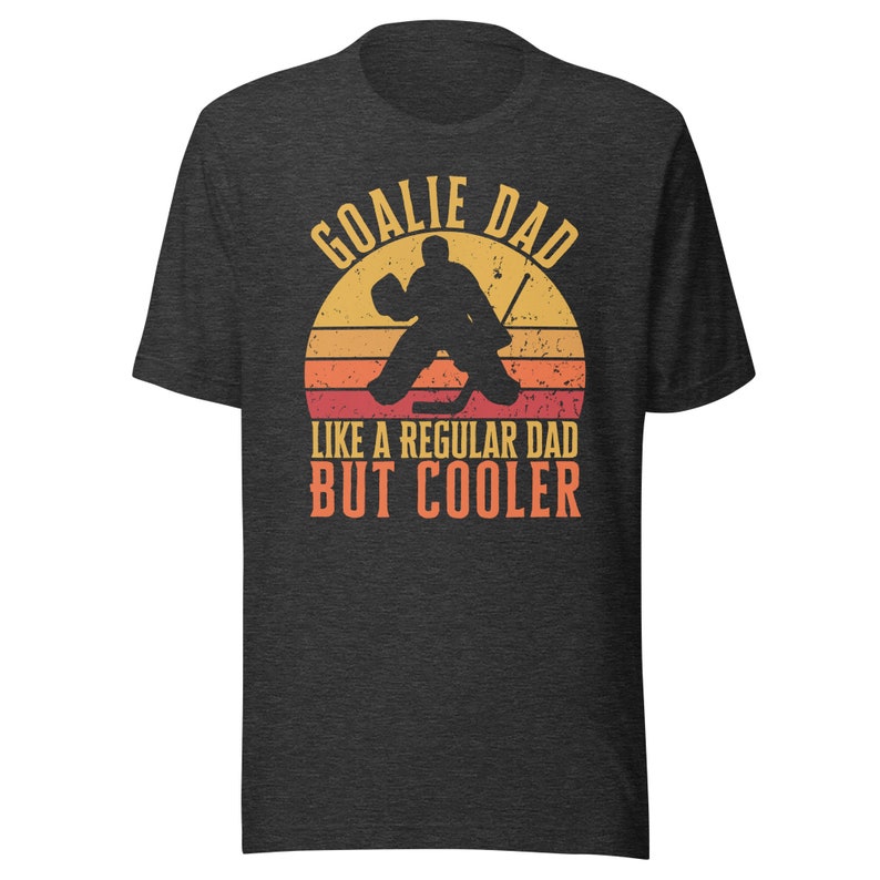 May include: A dark gray t-shirt with a vintage-style graphic of a hockey goalie in front of a sunset. The text reads "Goalie Dad Like a Regular Dad But Cooler".