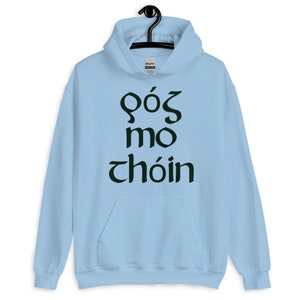 Puede incluir: Una sudadera con capucha azul claro con un bolsillo delantero y una capucha con cord&oacute;n. La sudadera con capucha presenta texto verde oscuro en una fuente estilizada. El texto dice "G&oacute;s mo ch&oacute;in". La sudadera con capucha est&aacute; colgada de una percha negra.