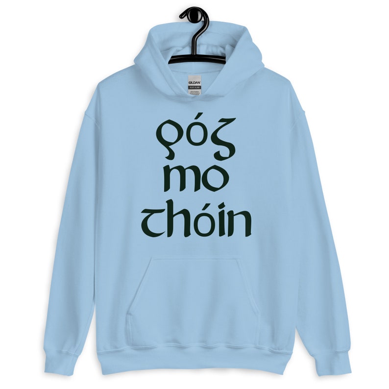 Puede incluir: Una sudadera con capucha azul claro con un bolsillo delantero y una capucha con cord&oacute;n. La sudadera con capucha presenta texto verde oscuro en una fuente estilizada. El texto dice "G&oacute;s mo ch&oacute;in". La sudadera con capucha est&aacute; colgada de una percha negra.