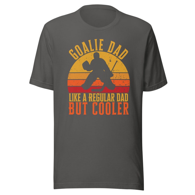 May include: A dark gray t-shirt with a retro-style graphic of a hockey goalie in front of a sunset. The text reads "Goalie Dad Like a Regular Dad But Cooler".