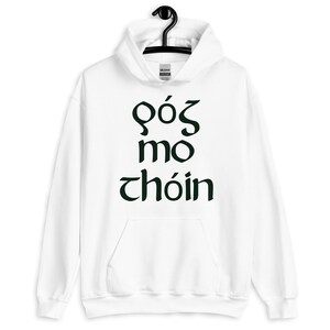 Puede incluir: Sudadera con capucha blanca con bolsillo delantero y cord&oacute;n. La sudadera presenta texto verde oscuro en una fuente estilizada. El texto dice "G&oacute;s mo ch&oacute;in". La sudadera est&aacute; colgada de una percha negra.