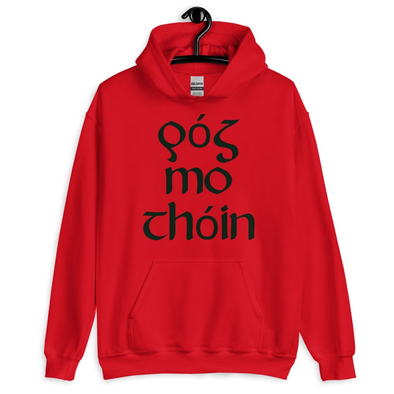 Puede incluir: Sudadera roja con capucha, bolsillo delantero y cord&oacute;n ajustable. La sudadera presenta texto verde oscuro en una fuente estilizada, posiblemente celta. La prenda est&aacute; colgada de una percha negra.