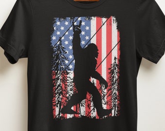 Puede incluir: Camiseta negra con una silueta de Bigfoot haciendo un gesto con la mano, sobre un diseño de bandera americana desgastada. El diseño incluye rayas rojas y blancas, estrellas azules y pinos.