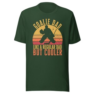 May include: Green t-shirt with a retro-style graphic of a hockey goalie in front of a sunset. The text reads "Goalie Dad Like a Regular Dad But Cooler".