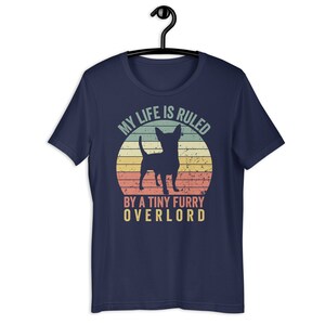 Op de afbeelding: Donkerblauw T-shirt met een vintage-stijl grafische afbeelding van een chihuahua-silhouet voor een zonsondergang. De tekst luidt "Mijn leven wordt geregeerd door een kleine harige overlord."