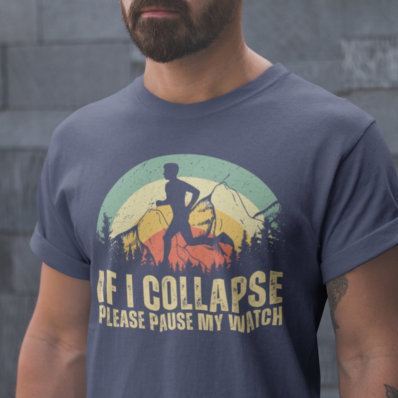 May include: A navy blue t-shirt with a vintage-style graphic of a runner in front of a mountain range and a sunset. The text "IF I COLLAPSE PLEASE PAUSE MY WATCH" is printed in yellow on the shirt.