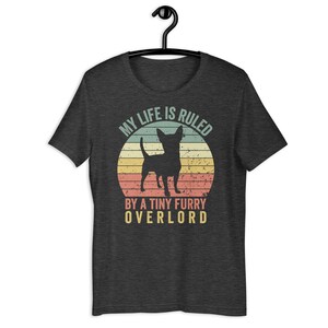 Op de afbeelding: Een donker grijs t-shirt met een vintage-stijl afbeelding van een chihuahua-silhouet voor een zonsondergang. De tekst luidt "Mijn leven wordt geregeerd door een kleine harige overlord."