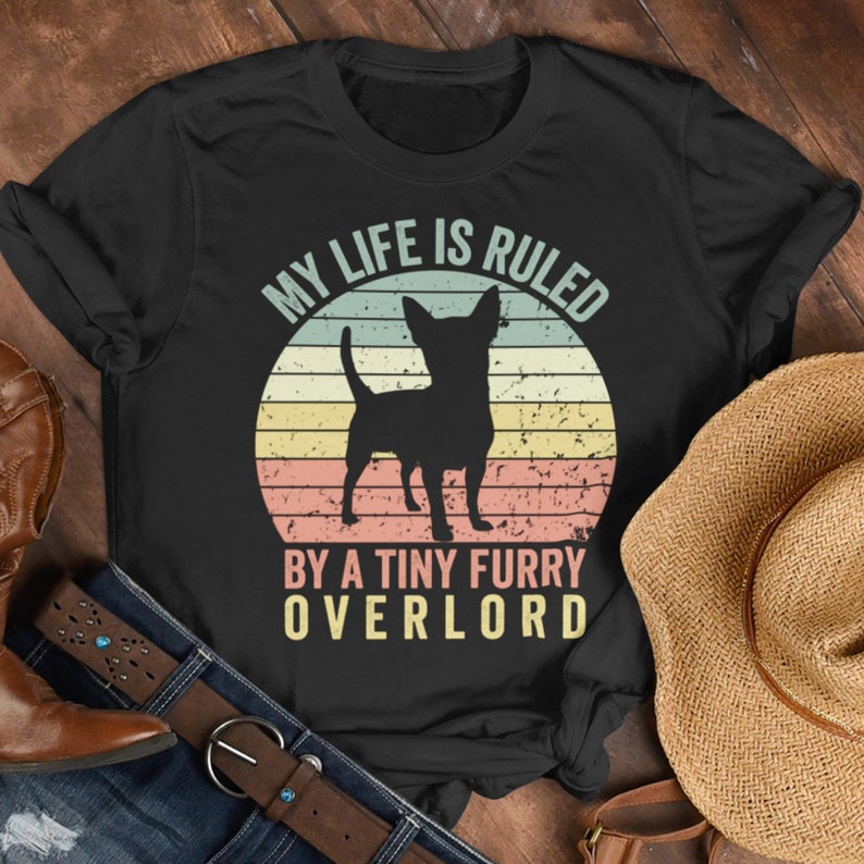 Op de afbeelding: Zwart T-shirt met een vintage-stijl afbeelding van een chihuahua en de tekst "My life is ruled by a tiny furry overlord".