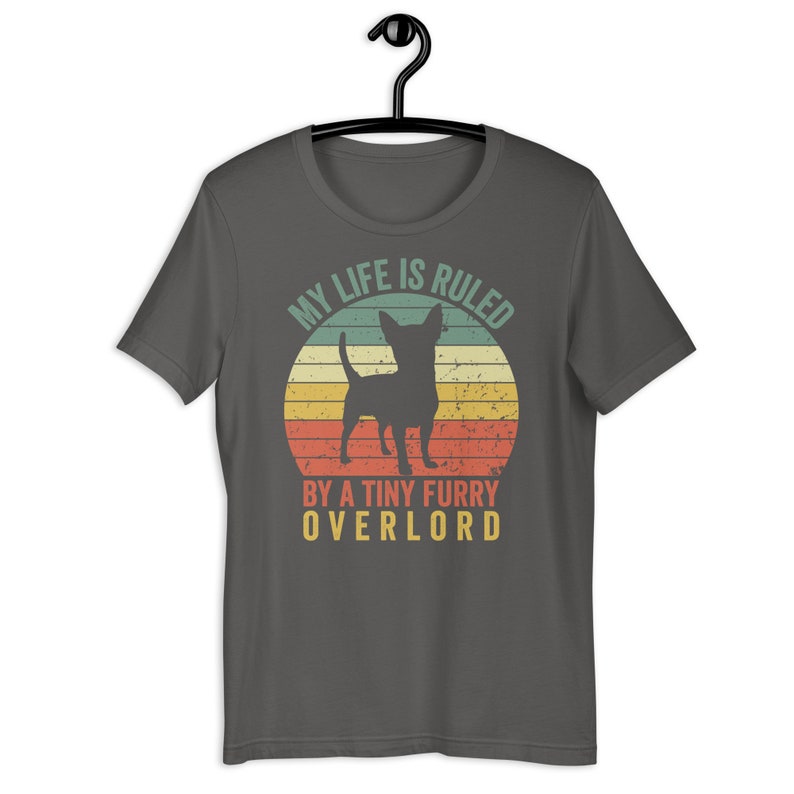 Op de afbeelding: Een donkergrijs T-shirt met een vintage-stijl grafische afbeelding van een chihuahua-silhouet voor een zonsondergang. De tekst luidt "Mijn leven wordt geregeerd door een kleine harige overlord."