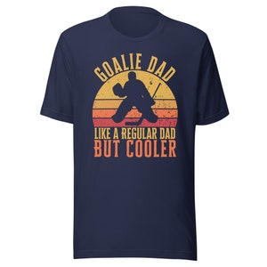 May include: Navy blue t-shirt with a vintage-style graphic of a hockey goalie in front of a sunset. The text reads "Goalie Dad Like a Regular Dad But Cooler".