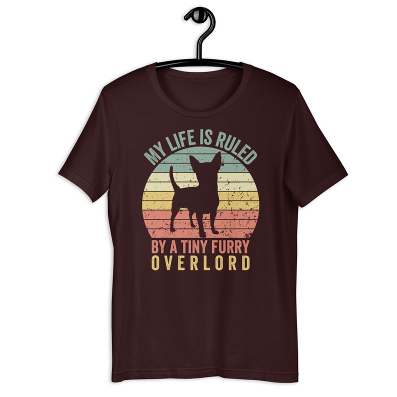 Op de afbeelding: Een maroon t-shirt met een vintage-stijl grafische afbeelding van een chihuahua-silhouet voor een zonsondergang. De tekst luidt "Mijn leven wordt geregeerd door een kleine harige overlord."