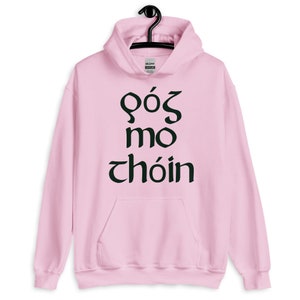 Puede incluir: Una sudadera con capucha rosa claro con un bolsillo delantero y una capucha con cord&oacute;n. La sudadera con capucha presenta texto negro en una fuente estilizada. El texto dice "GOS MO CHOIN". La sudadera con capucha est&aacute; colgada de una percha negra.