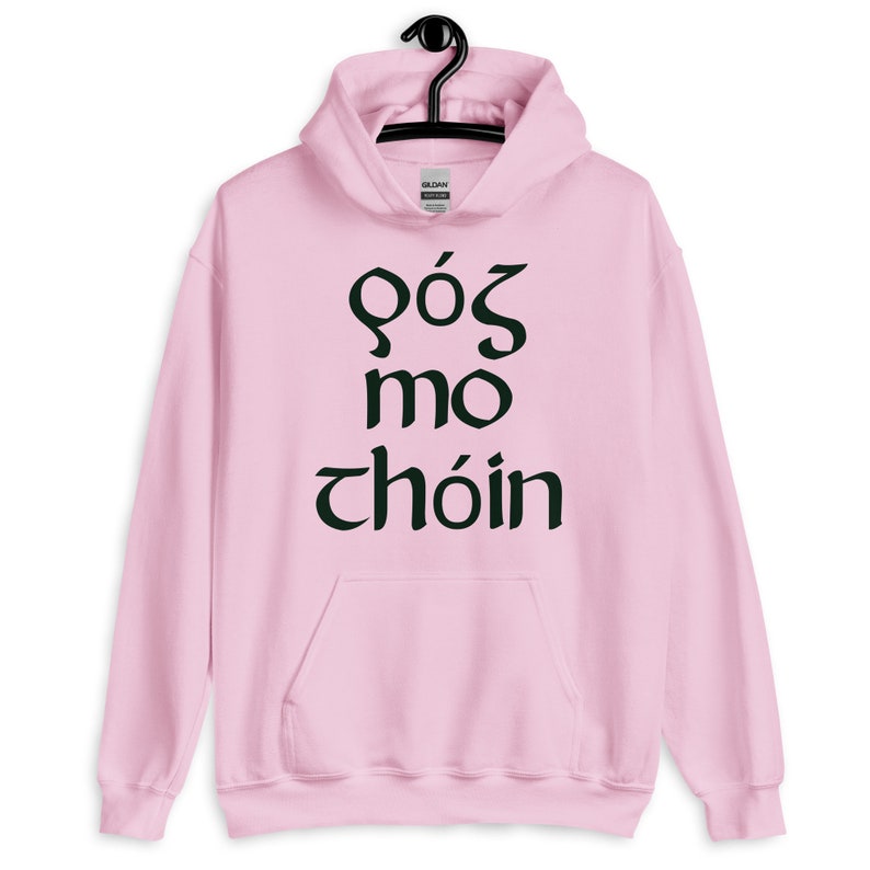 Puede incluir: Una sudadera con capucha rosa claro con un bolsillo delantero y una capucha con cord&oacute;n. La sudadera con capucha presenta texto negro en una fuente estilizada. El texto dice "GOS MO CHOIN". La sudadera con capucha est&aacute; colgada de una percha negra.