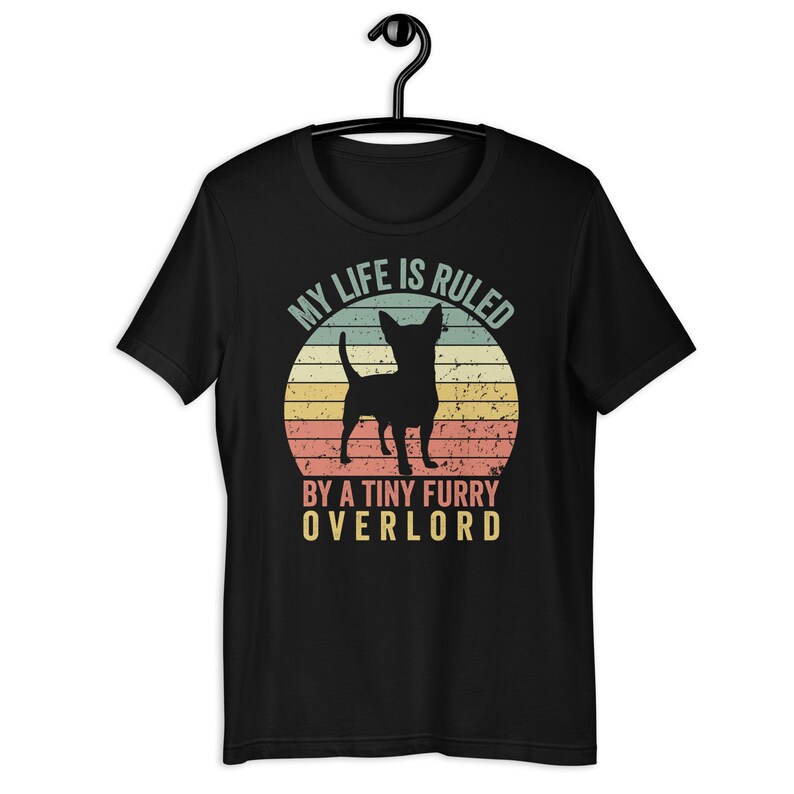 Op de afbeelding: Zwart T-shirt met een vintage-stijl grafische afbeelding van een chihuahua-silhouet voor een zonsondergang. De tekst luidt "Mijn leven wordt geregeerd door een kleine harige overlord."