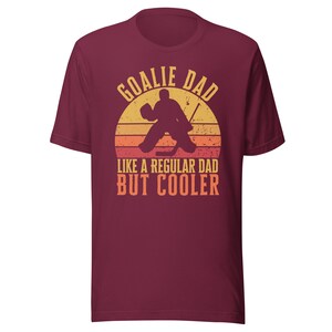 May include: Burgundy t-shirt with a retro-style graphic of a hockey goalie in front of a sunset. The text reads "Goalie Dad Like a Regular Dad But Cooler."