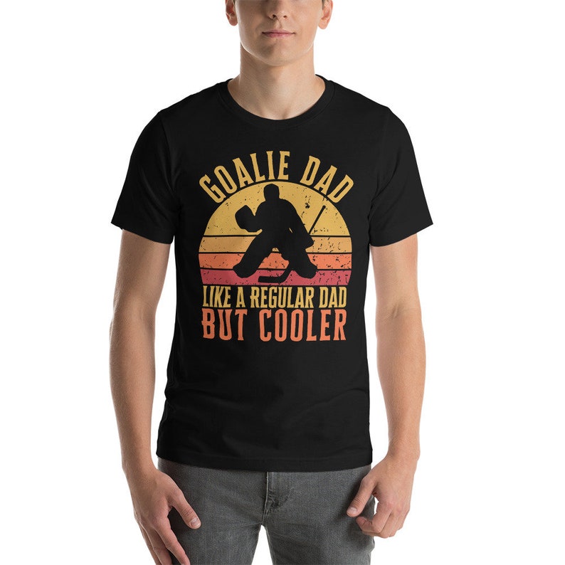 May include: Black t-shirt with a vintage-style graphic of a hockey goalie in front of a sunset. The text reads "Goalie Dad Like a Regular Dad But Cooler."