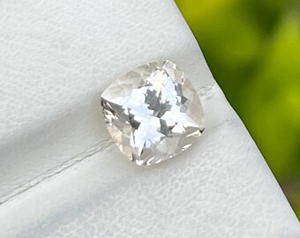 May include: A clear, cushion-cut gemstone with multiple facets, reflecting light. The gem is set against a white background, with a blurred green backdrop. The gemstone's shape is square with rounded corners.