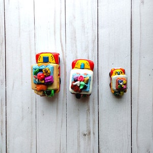 May include: Three colorful toy trucks with painted details. The largest truck is red, yellow, and green with a bed full of colorful fruit and vegetables. The middle truck is red, yellow, and white with a bed full of colorful flowers. The smallest truck is red, yellow, and white with a bed full of colorful flowers.