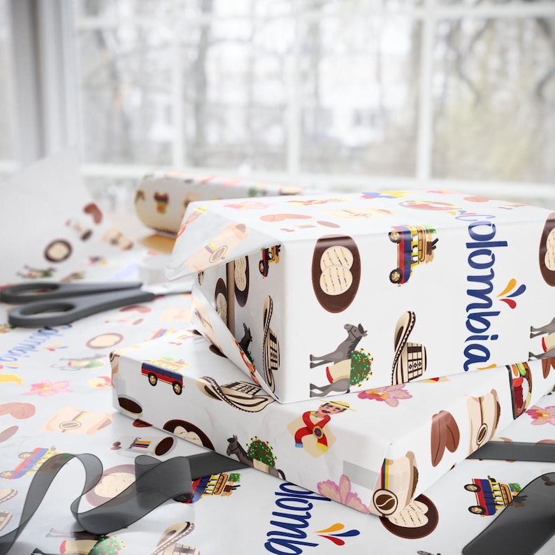 May include: Two gift boxes wrapped in white paper printed with colorful Colombian-themed illustrations. The word "Colombia" is printed in blue on the side of the boxes. A pair of scissors and a roll of black ribbon are on the table.