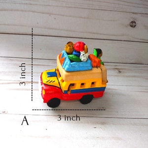 May include: A small, colorful, hand-painted ceramic toy bus with a yellow body, red front, and blue accents. The bus is carrying a variety of colorful fruits and vegetables on top. The bus is 3 inches long and 3 inches tall.