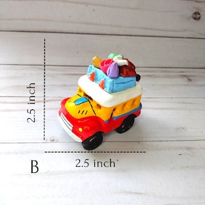 May include: A small, colorful, ceramic toy car with a red body, yellow sides, and a white roof. The car is loaded with luggage on top, including a blue suitcase, a pink suitcase, and a brown bag. The car is 2.5 inches wide and 2.5 inches tall.
