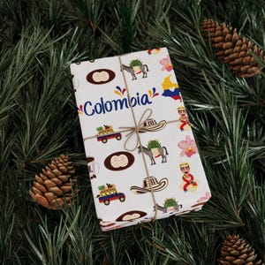 May include: A wrapped gift with a white background featuring colorful illustrations of Colombia. The design includes images of donkeys, hats, coffee cups, and the country's flag. The word "Colombia" is written in blue script.