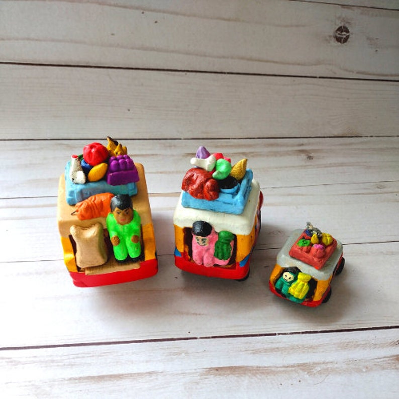 May include: Three colorful toy trucks with various fruits and vegetables on top. The trucks are made of clay and have a hand-painted finish. The trucks are on a white wooden surface.