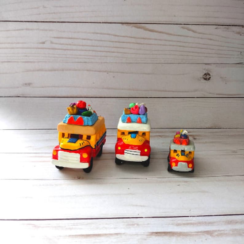 May include: Three colorful toy buses made of clay with different designs and sizes. The largest bus is red, yellow, and blue with a white front and a variety of colorful decorations on top. The middle bus is similar in design but smaller. The smallest bus is red, yellow, and white with a white front and a simple design.